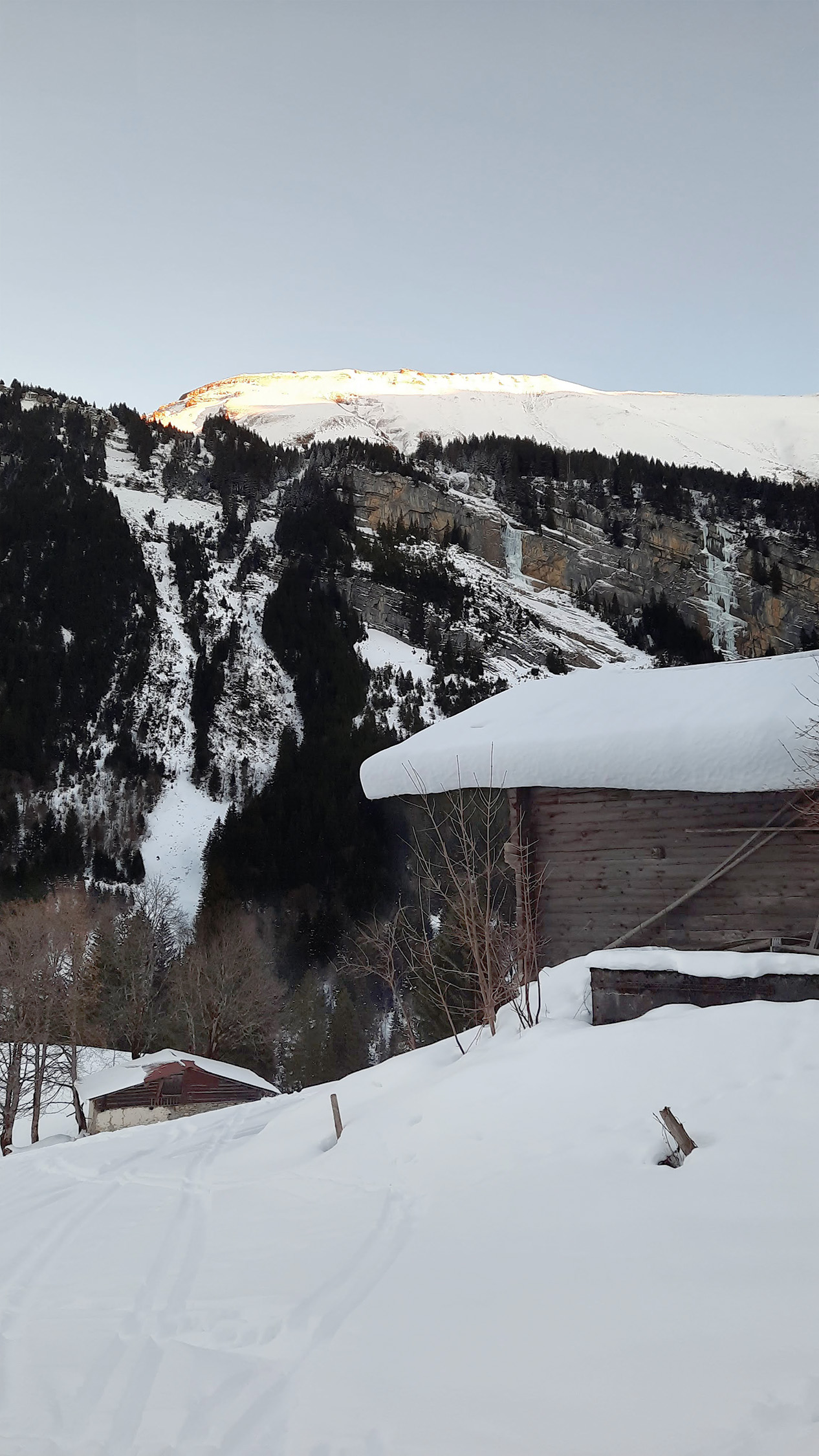 A snowy mountainside lit by evening sun, with a wooden chalet in the foreground